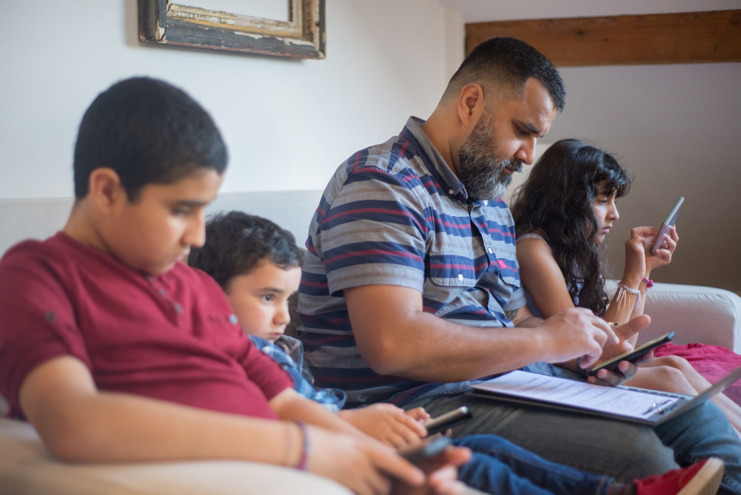Padre e hijos utilizando diversos dispositivos electrónicos en el sofá de casa, ilustrando la protección de red para toda la familia.