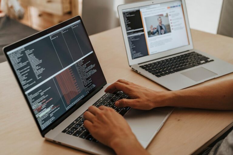 Desarrollador trabajando en un entorno de doble portátil; en primer plano escribe código en un editor de texto oscuro para un script de scraping, mientras en el portátil de fondo se visualiza la página web objetivo.