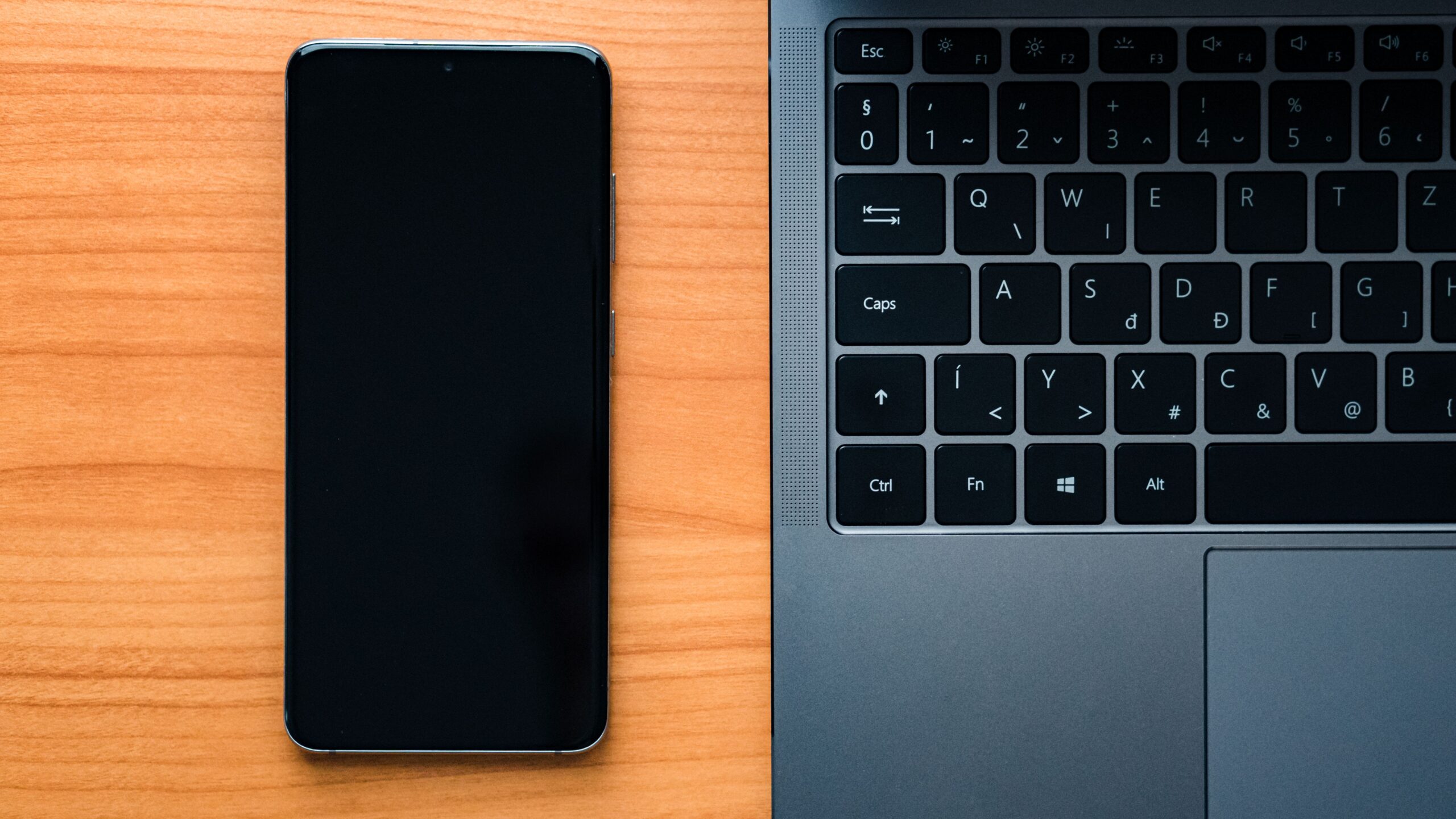 Vista cenital de un ordenador portátil y un smartphone sobre un escritorio de madera, representando la conexión entre distintos dispositivos.
