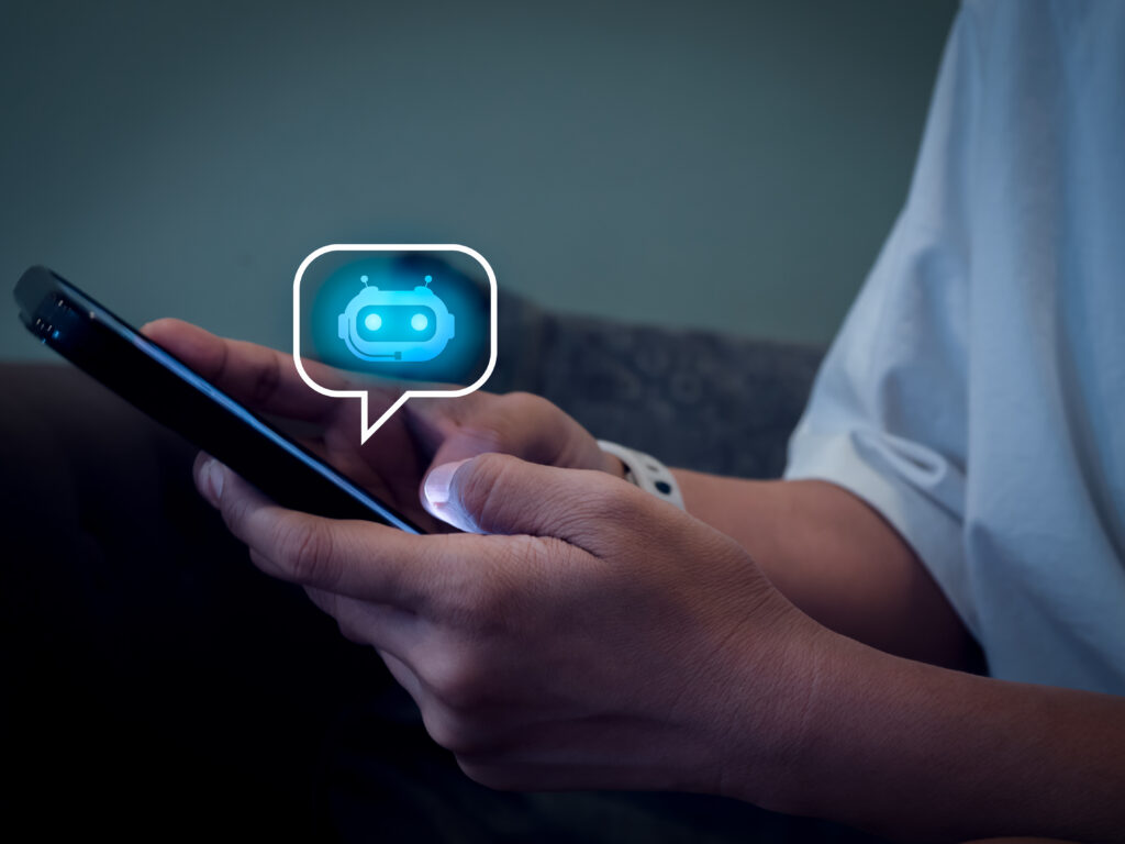 Icono de robot inteligente brillante dentro de una burbuja de habla en la pantalla de un teléfono móvil sostenido en las manos, representando el concepto de chatbot y asistente de conversación con IA