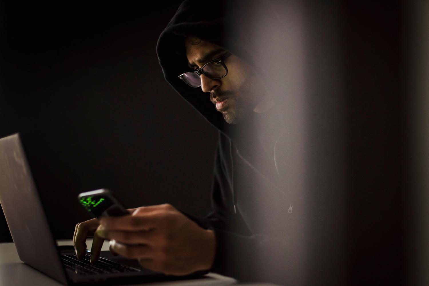 Imagen atmosférica de un hacker con capucha utilizando un portátil y un móvil, ilustrando los riesgos de ciberseguridad.