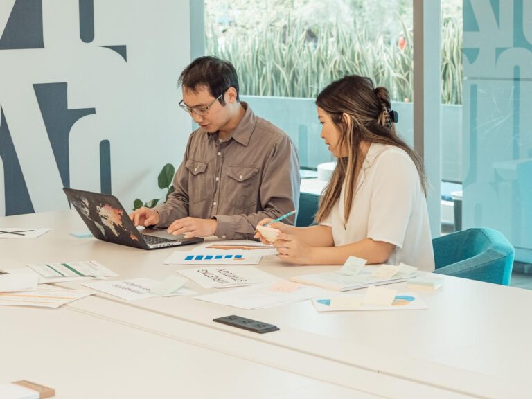 Dos colegas colaborando en una oficina moderna sobre un portátil y documentos, representando el trabajo en equipo y la cocreación.