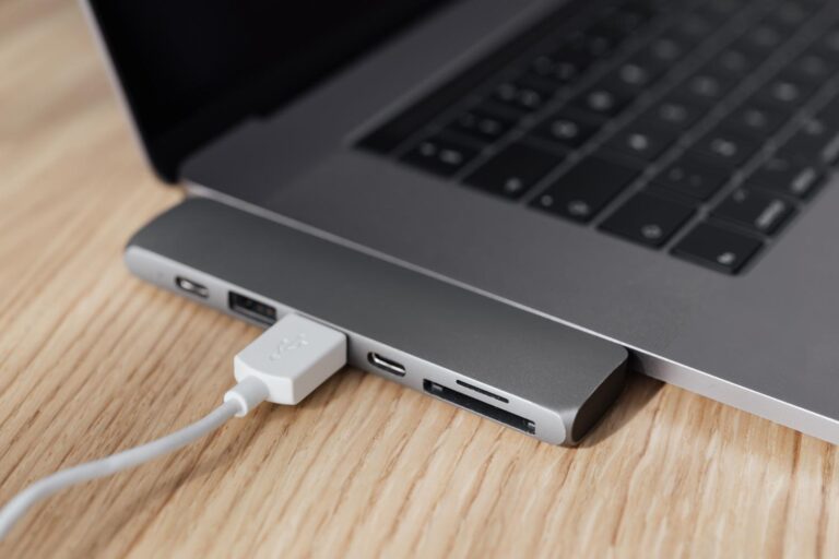 Primer plano de un cable USB blanco conectado a un hub multipuerto en el lateral de un ordenador portátil plateado sobre una mesa de madera, ilustrando la conexión de hardware periférico.