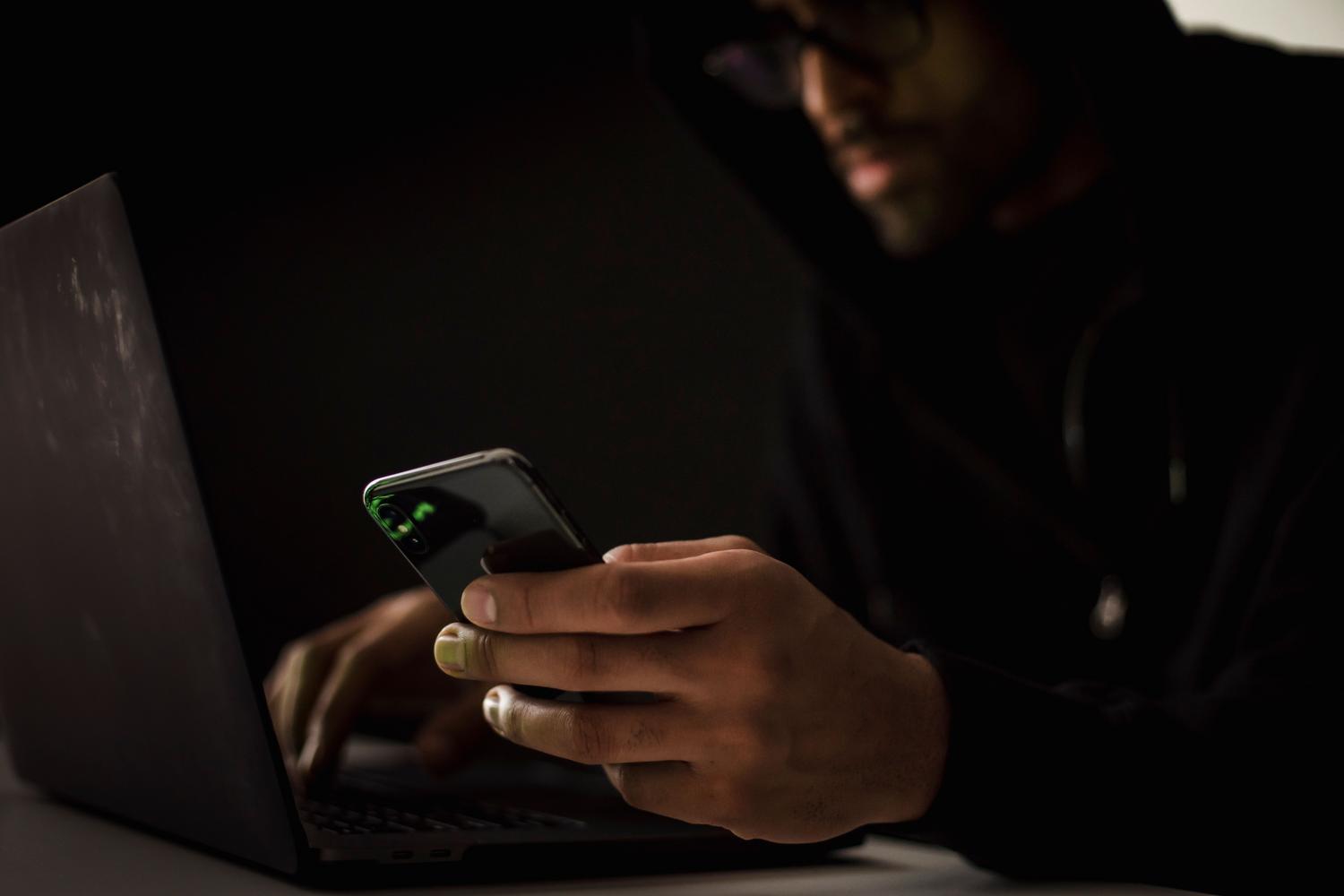 Persona utilizando un móvil y un portátil en un entorno oscuro, como representación de una amenaza digital.