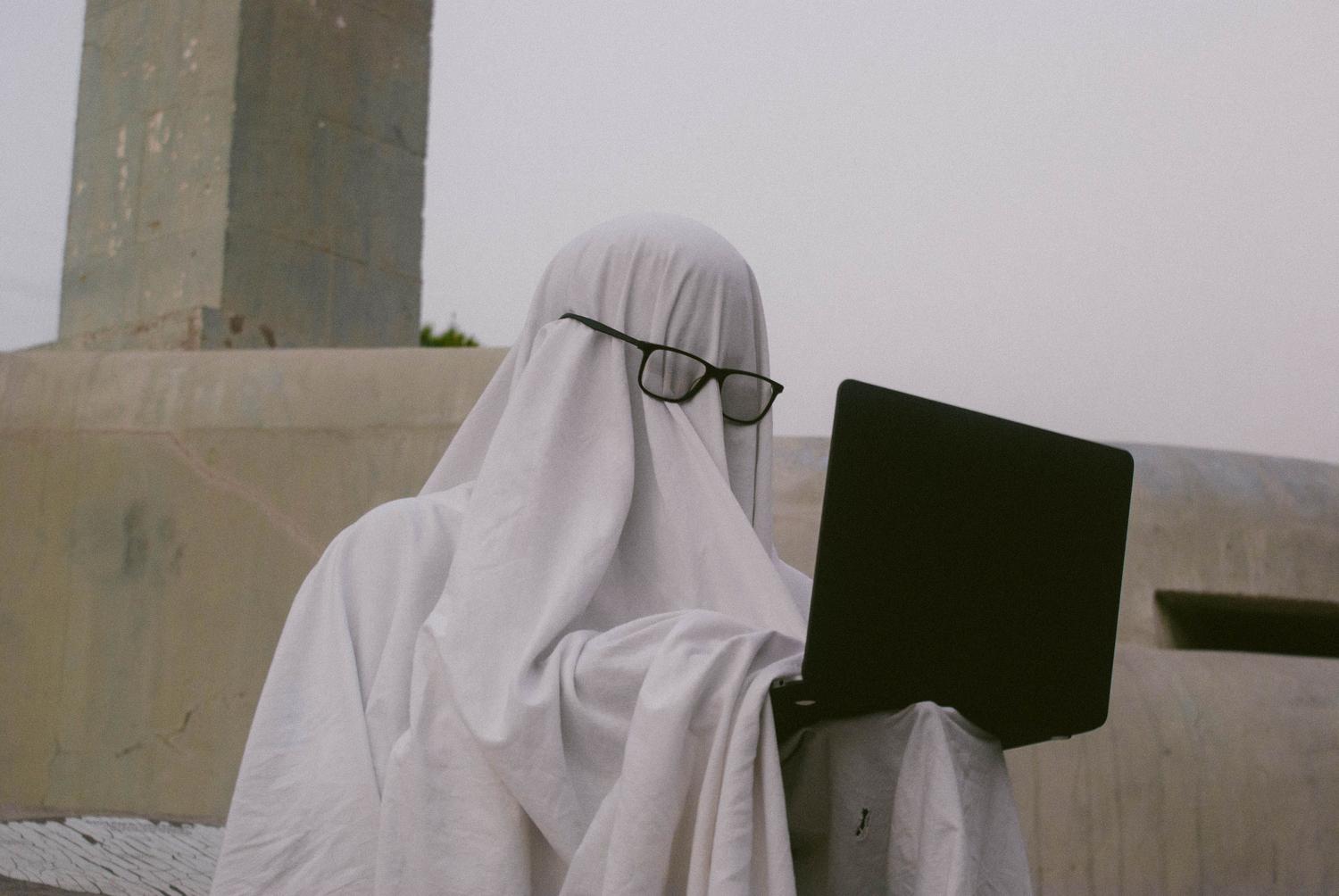 Persona disfrazada de fantasma usando un portátil, ideal para ilustrar bromas de Halloween.