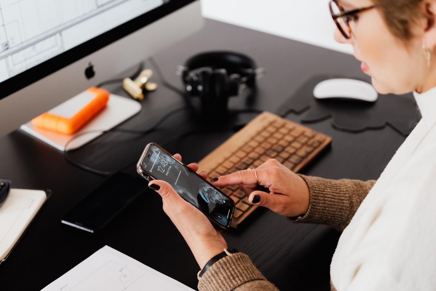 Mujer utilizando su teléfono móvil mientras trabaja en el ordenador, ilustrando la multitarea entre dispositivos.