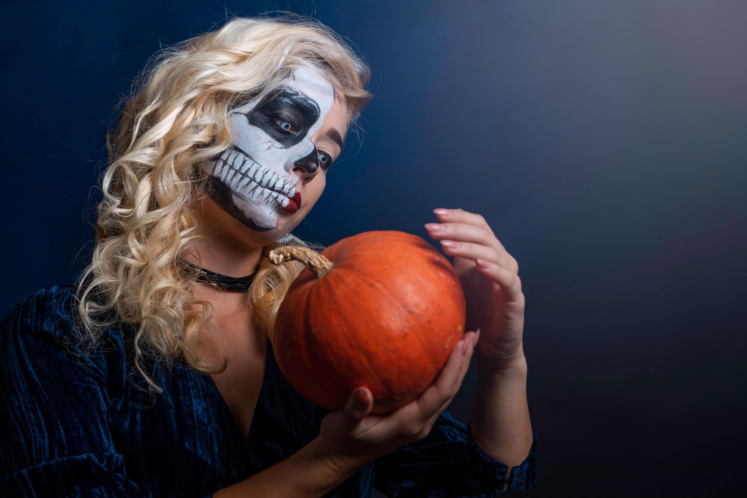 Künstlerisches Porträt einer Frau mit Halbschädel-Make-up, die einen Halloween-Kürbis hält.