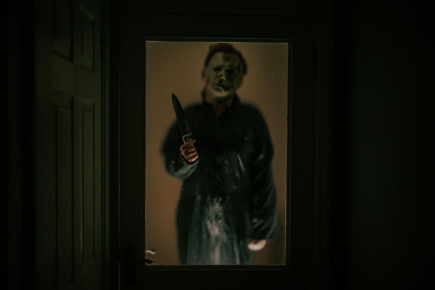Figura enmascarada acechando frente a una puerta tenuemente iluminada durante la noche de Halloween.