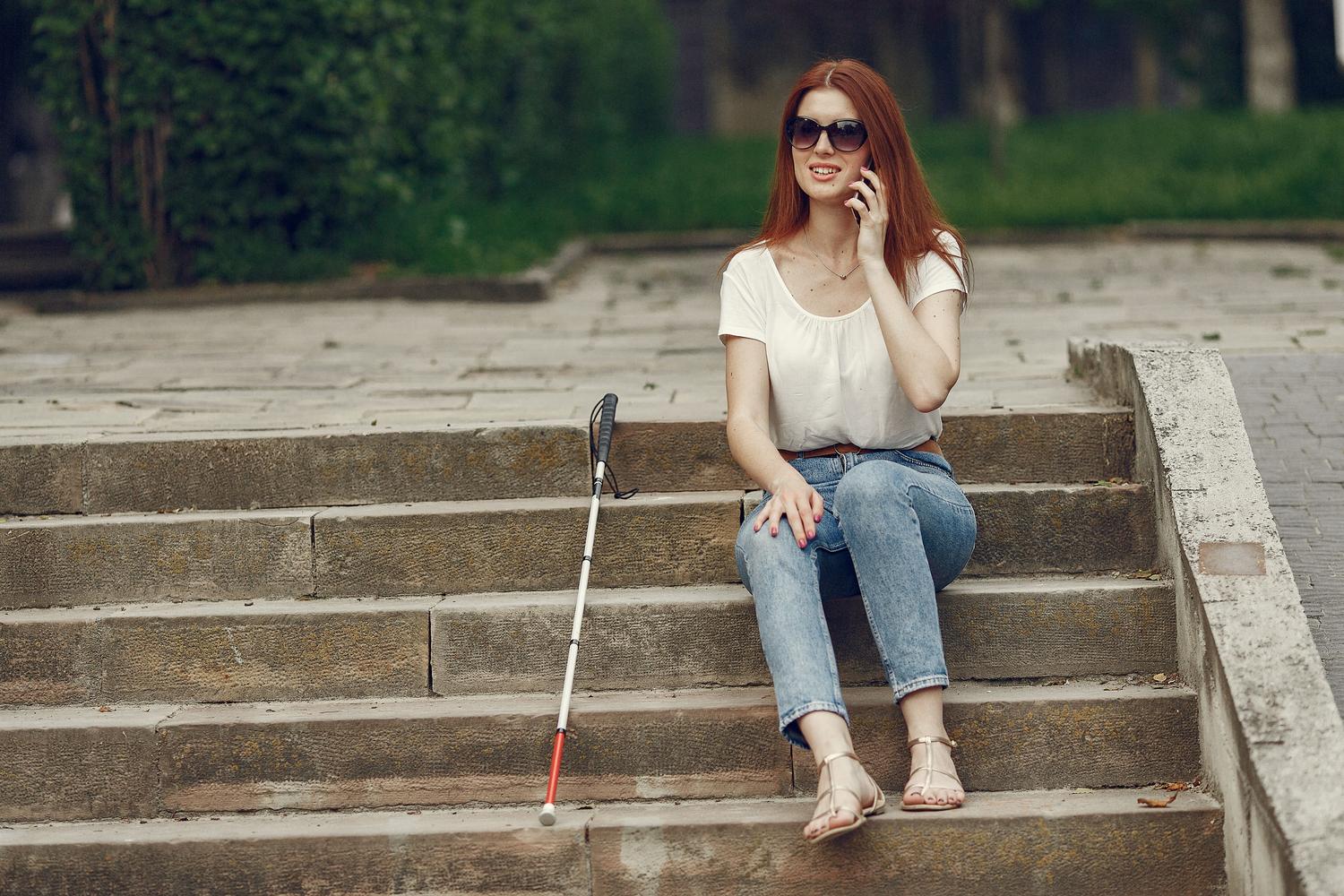 Mujer con discapacidad visual sentada al aire libre utilizando su teléfono móvil para interactuar con el entorno. Mujer con discapacidad visual sentada al aire libre utilizando su teléfono móvil para interactuar con el entorno.