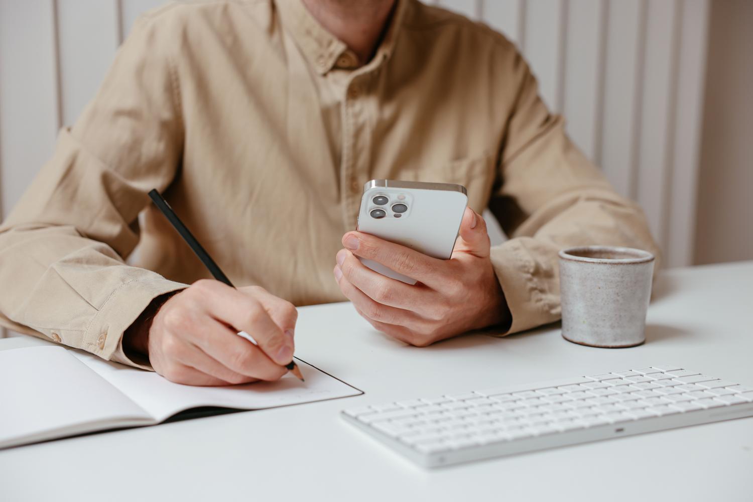 Persona tomando notas en un cuaderno mientras consulta información en su teléfono móvil.
