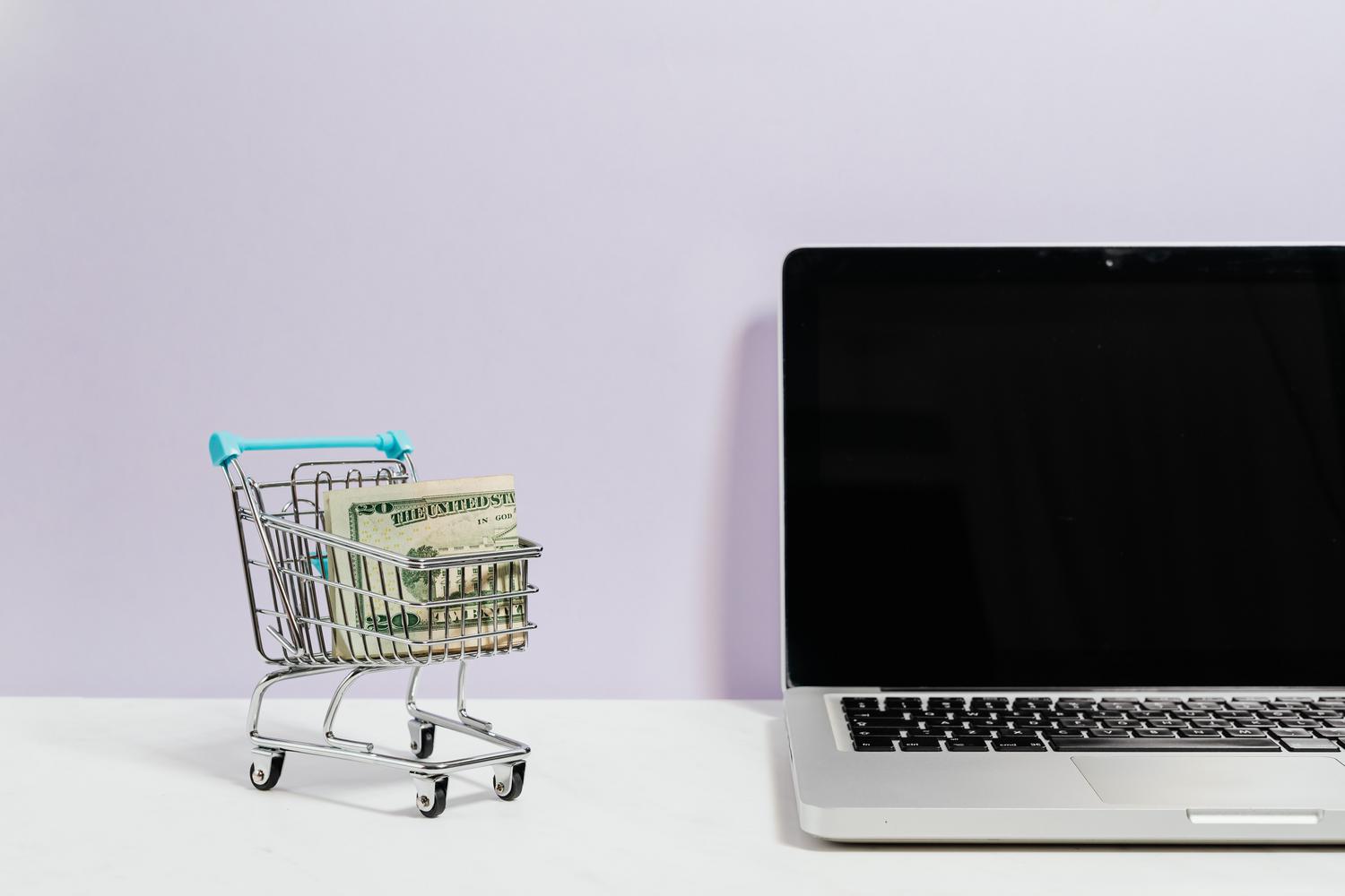 Mini carrito de compras con dinero junto a un ordenador portátil, representando el rastreo de stock y precios en e-commerce.