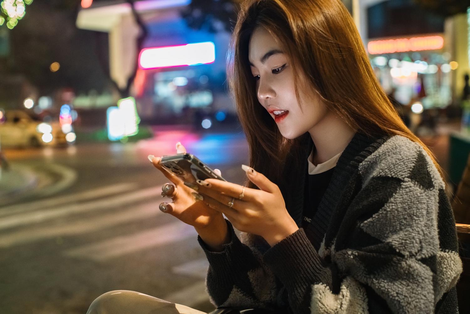Una joven utiliza su teléfono móvil en una animada calle de una ciudad asiática durante la noche, reflejando el estilo de vida digital urbano.