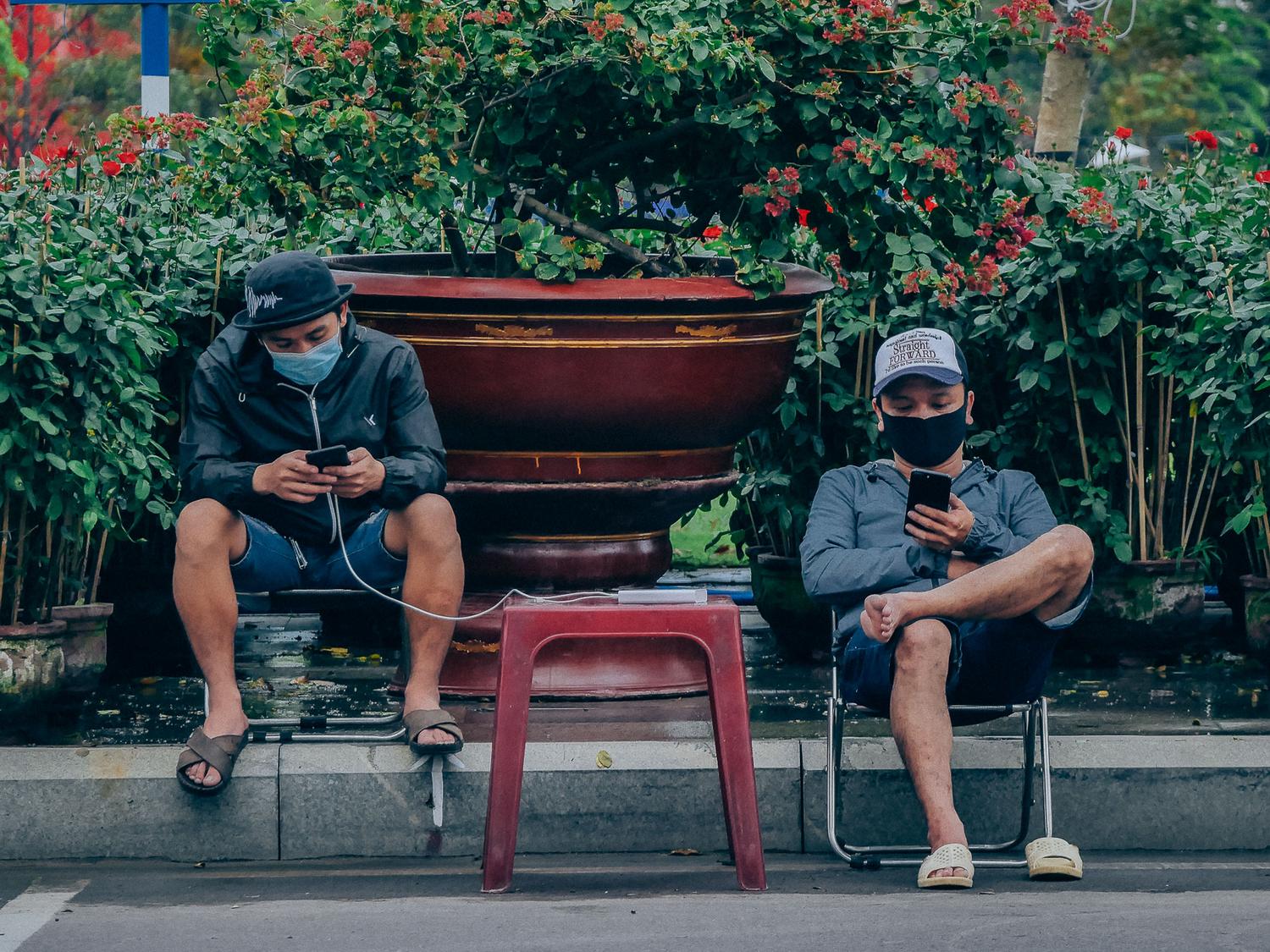 Dos hombres sentados al aire libre en Vietnam utilizando sus smartphones, ejemplificando la adopción masiva de servicios móviles en el sudeste asiático.