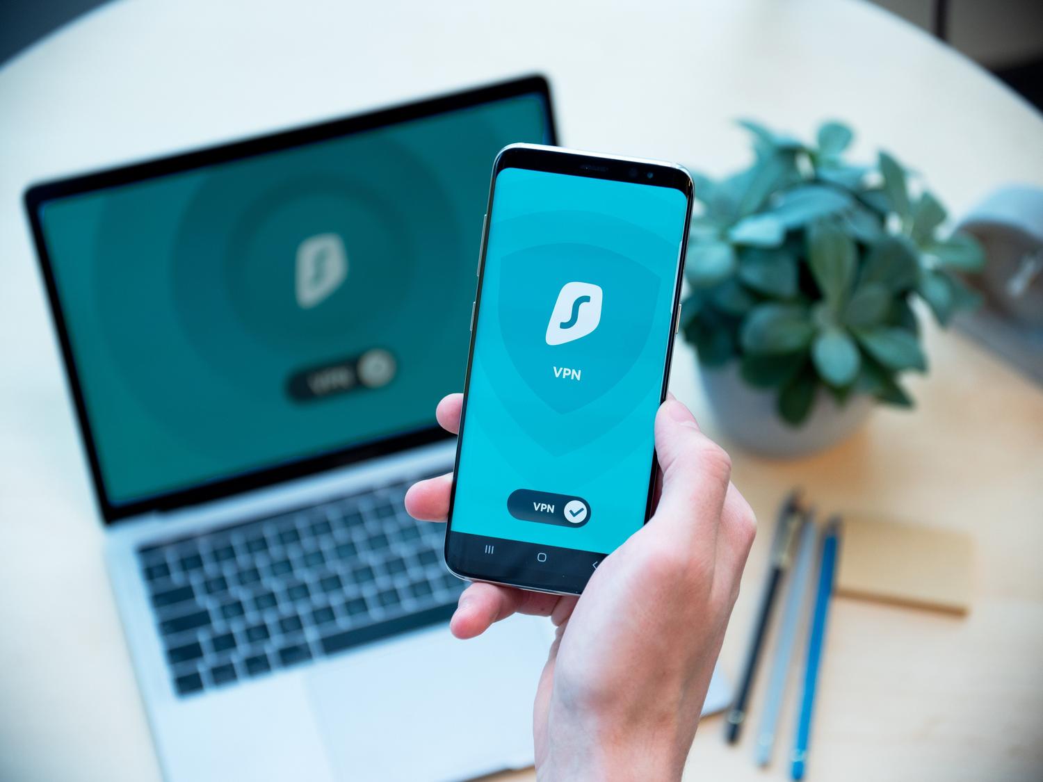 Mano sosteniendo un smartphone con una aplicación de seguridad frente a un portátil, ilustrando la verificación en dos pasos y la gestión de dispositivos para la seguridad del correo electrónico.