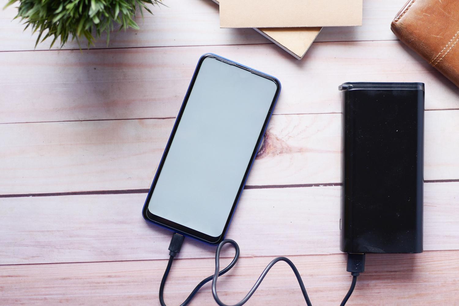 Smartphone conectado a una batería externa o powerbank sobre un escritorio de madera, como solución temporal al drenaje de batería.