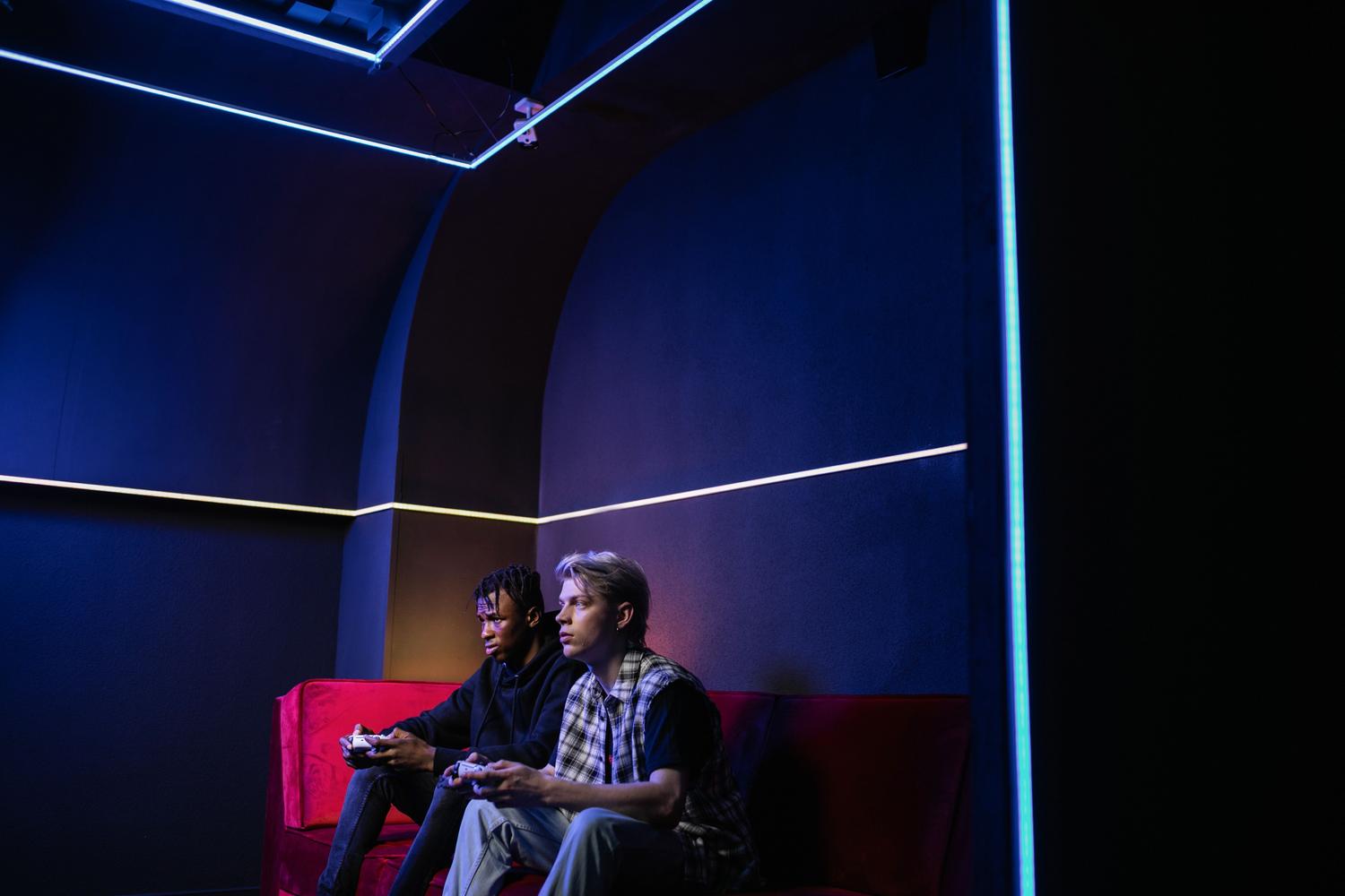 Dos amigos jugando videojuegos en un salón con luces de neón y ambiente oscuro, representando la experiencia social de jugar en cooperativo.