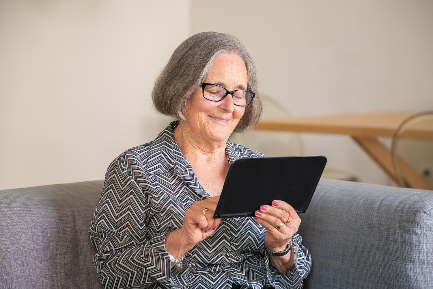 Mujer mayor sonriendo mientras utiliza una tablet en su hogar, representando la accesibilidad de la tecnología en el cuidado familiar. Mujer mayor sonriendo mientras utiliza una tablet en su hogar, representando la accesibilidad de la tecnología en el cuidado familiar.