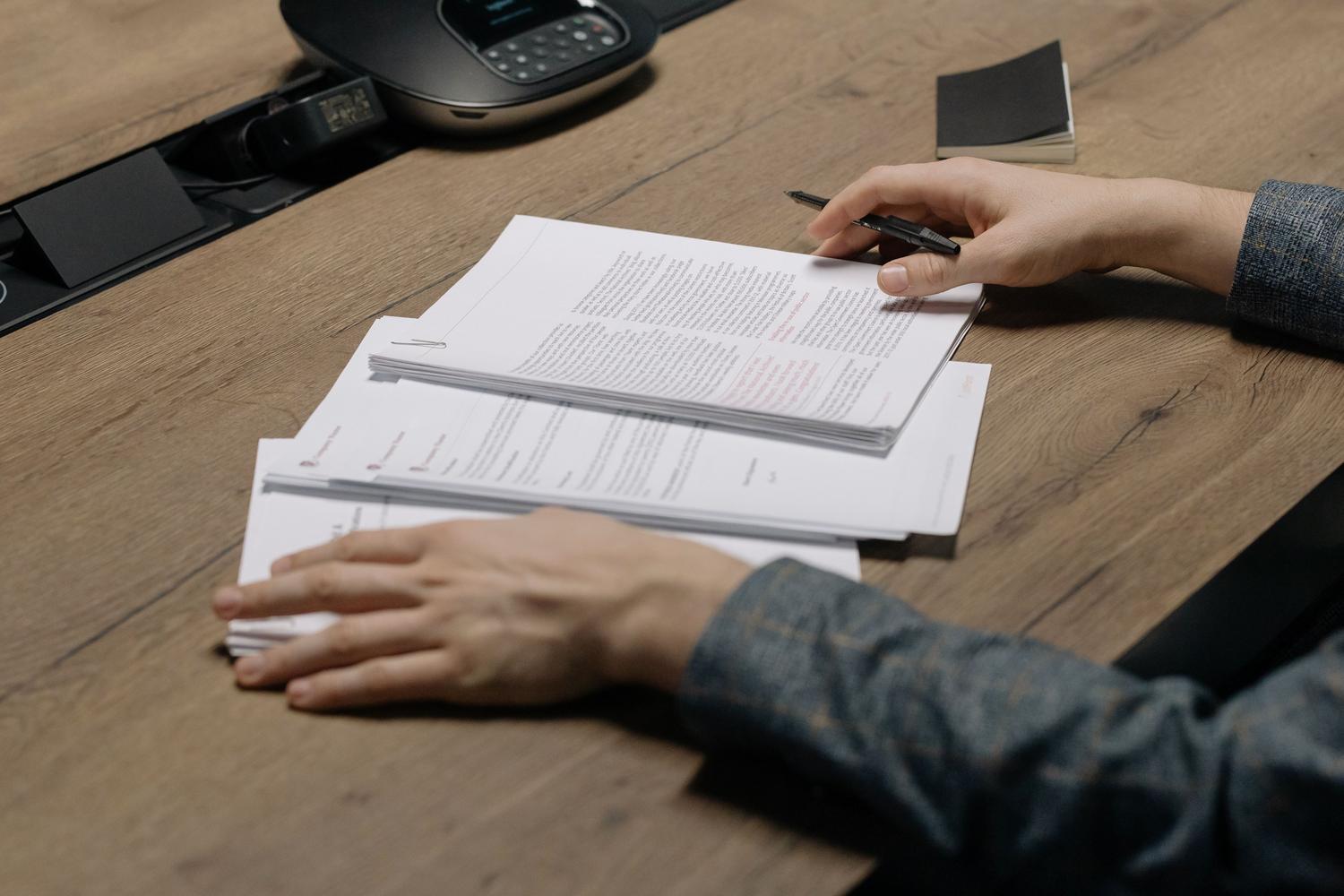 Primer plano de una persona revisando documentos con un bolígrafo, representando la verificación de los importes y fechas corregidos.