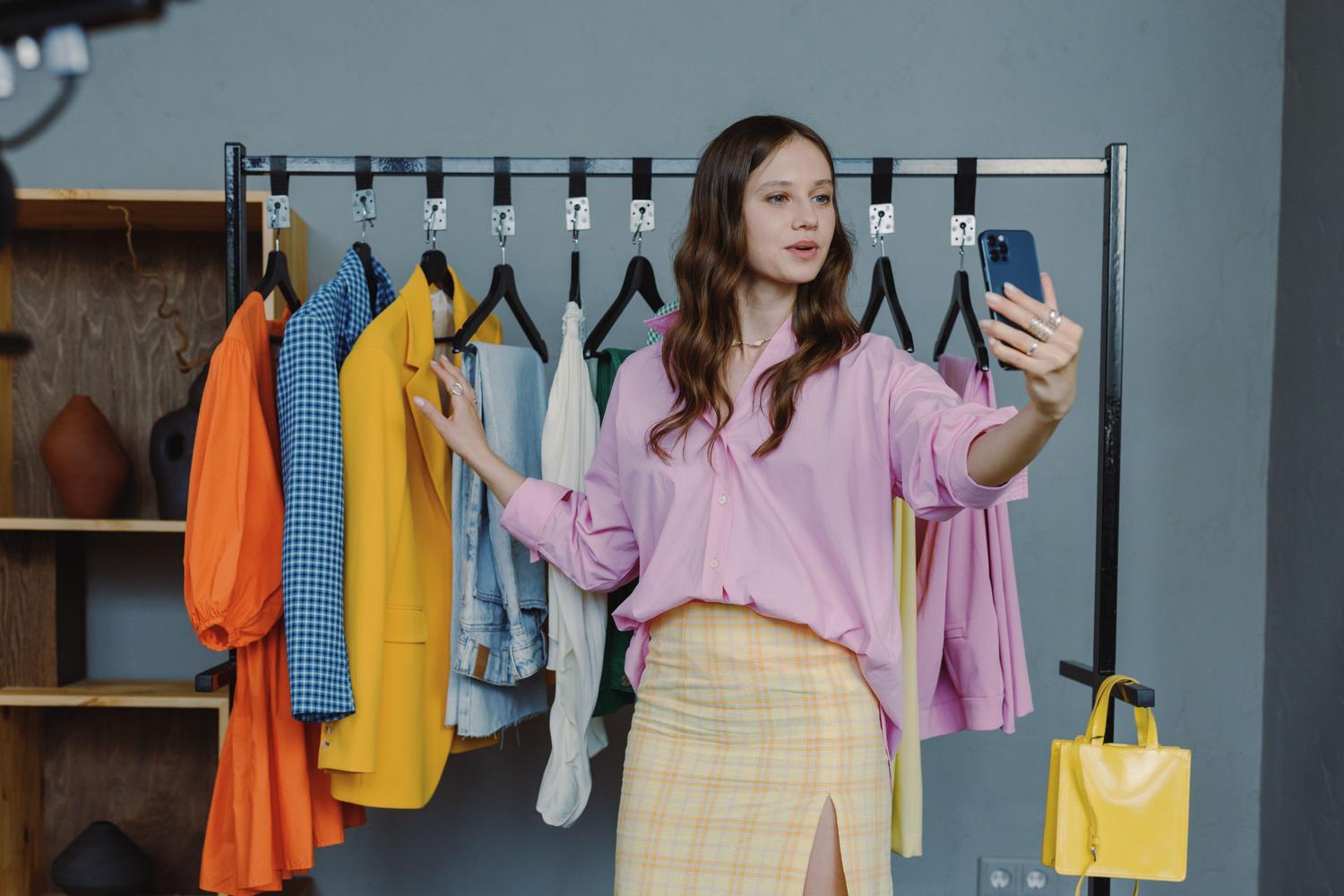 Woman using a smartphone to document her outfits in front of a colorful clothing rack, representing the 'Digital Wardrobe' concept.